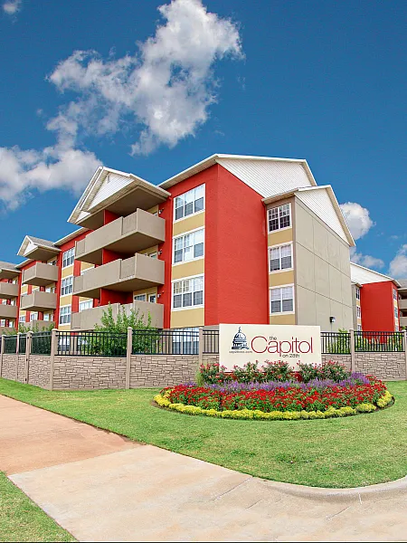 Modern apartment building with bold red and beige facade, featuring spacious balconies and surrounded by vibrant landscaping under a clear blue sky.