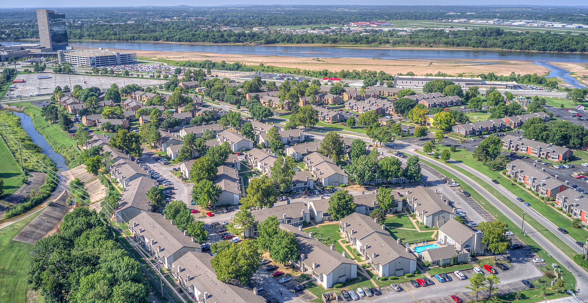 Aerial view of a serene apartment complex surrounded by lush greenery, with well-organized buildings and a nearby river, offering a peaceful living environment.