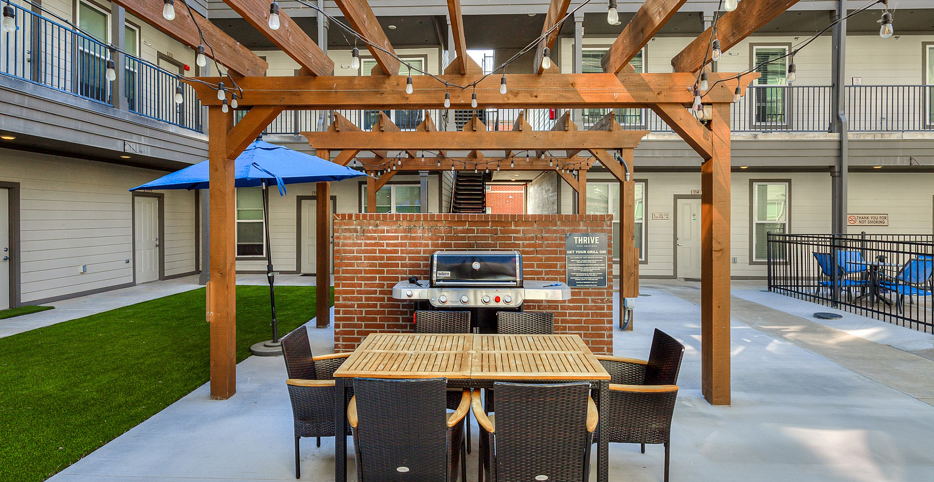 Charming apartment courtyard featuring a stylish pergola with string lights, inviting outdoor dining area, and modern BBQ grill, perfect for entertaining.