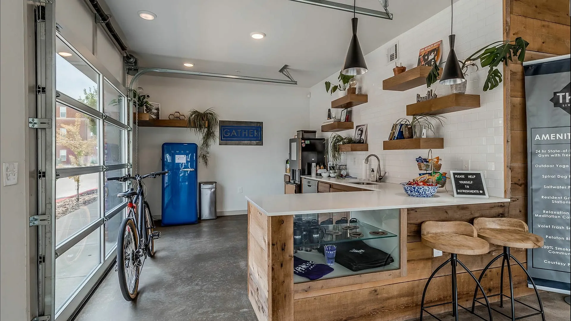 Chic modern lounge area with open garage-style doors, a vibrant blue fridge, and stylish wooden shelving. Inviting bar counter with cozy seating and lush greenery adds a refreshing touch.