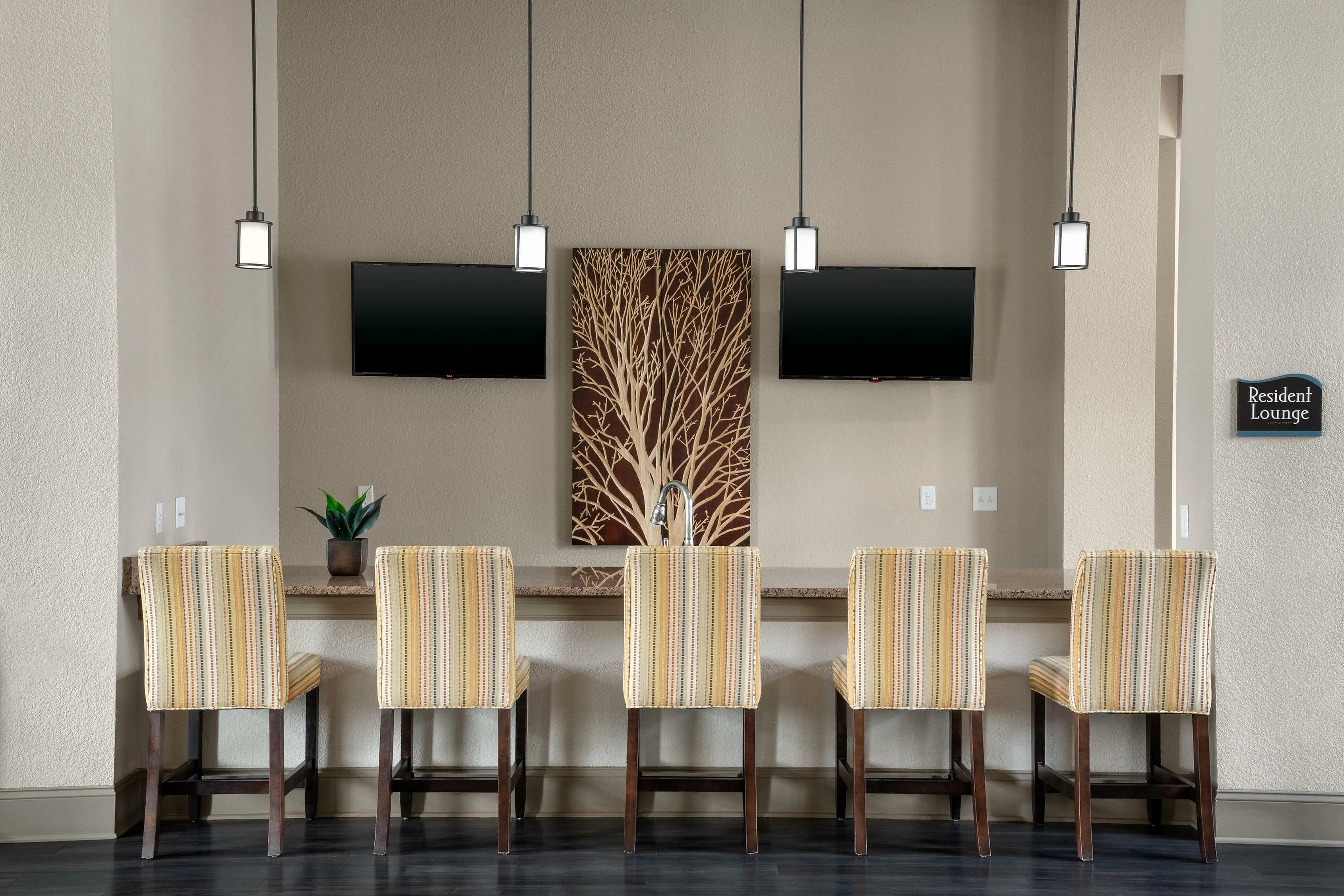 Chic resident lounge with a row of stylish chairs, dual mounted TVs, elegant pendant lighting, and modern decor.