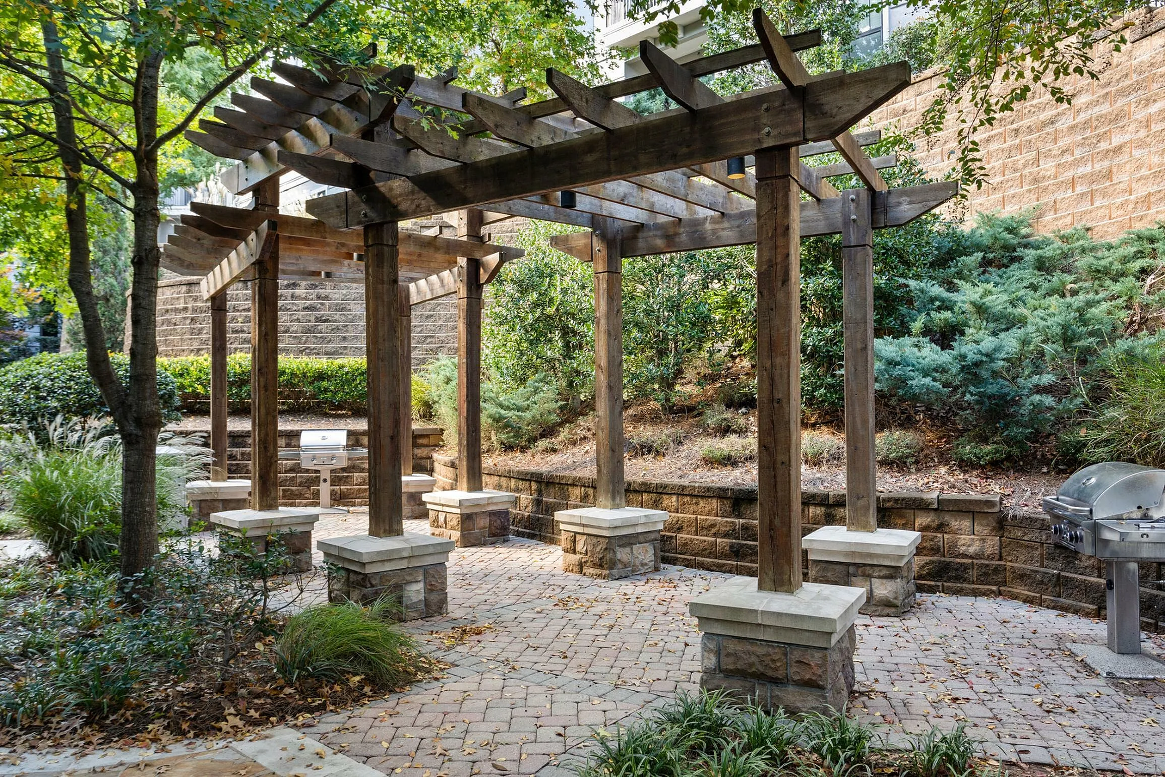 Inviting outdoor patio area with wooden pergola and built-in grill set amidst lush greenery.