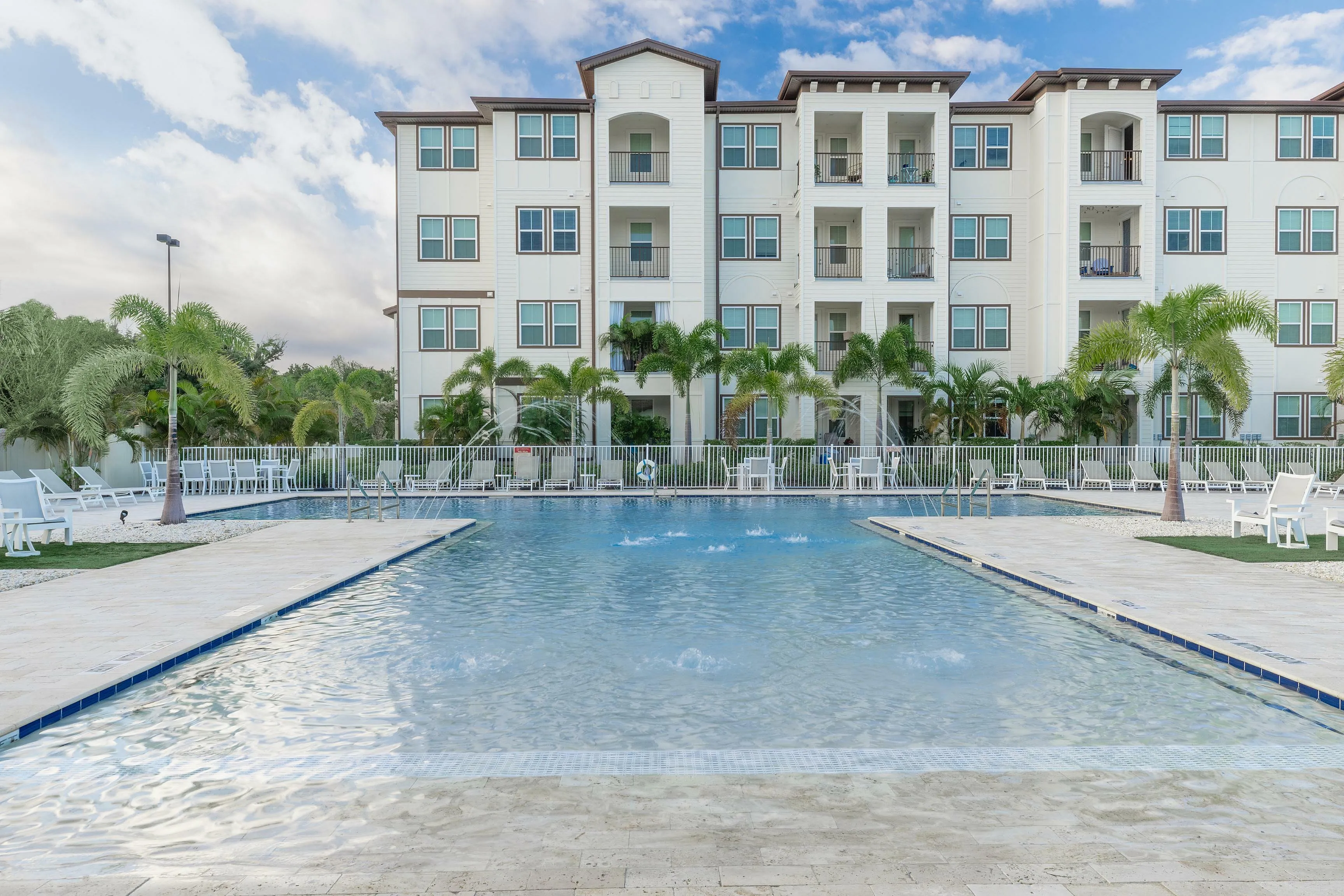 Elegant apartment complex with a shimmering pool surrounded by lush palm trees and seating areas.