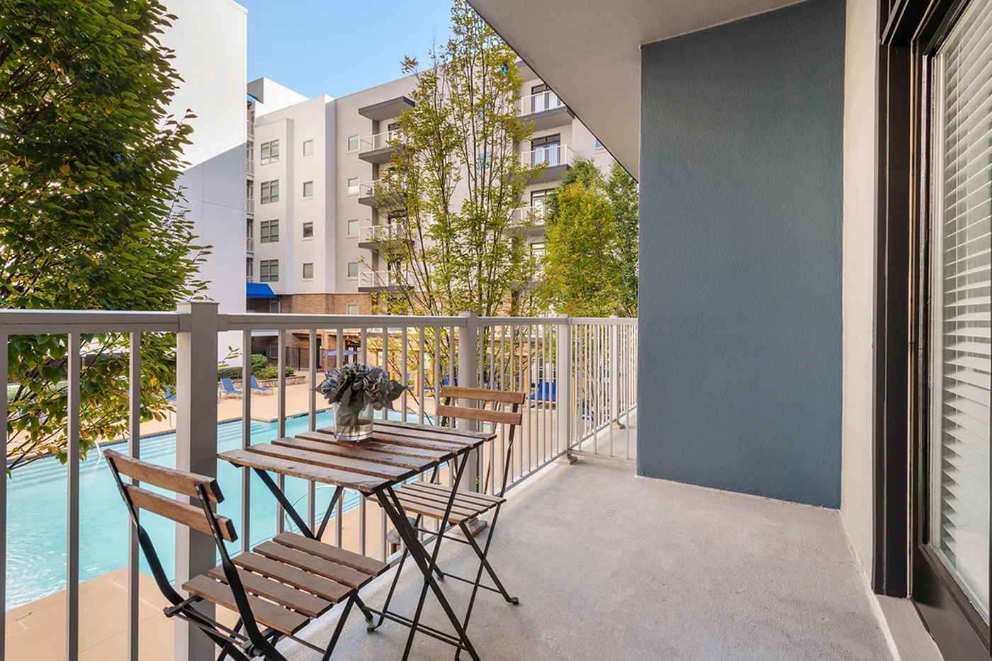 Charming balcony view from a modern apartment overlooking a sunlit swimming pool and lush greenery.