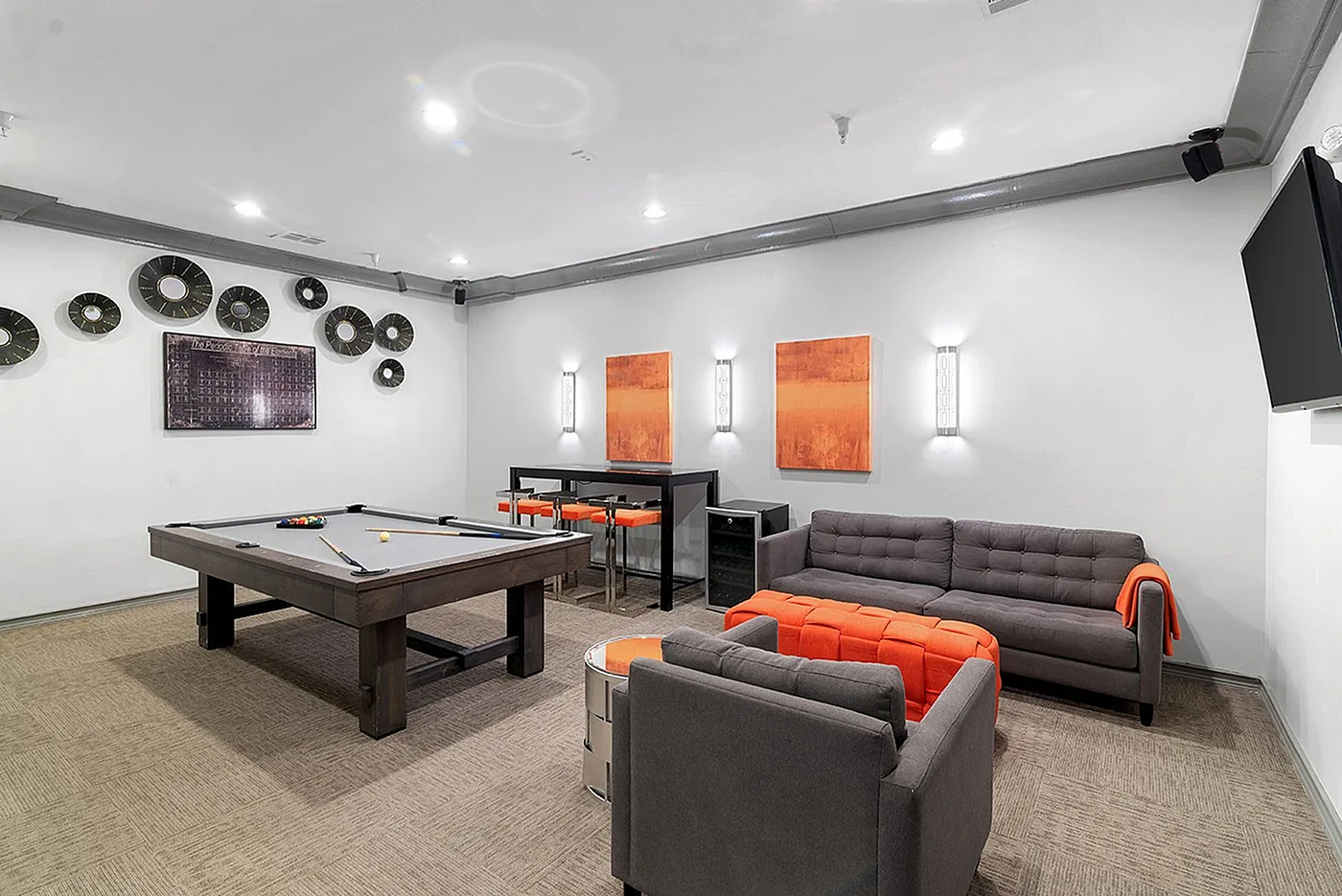 Modern entertainment room featuring a sleek pool table, stylish grey sofas with vibrant orange accents, and contemporary wall art and decor.