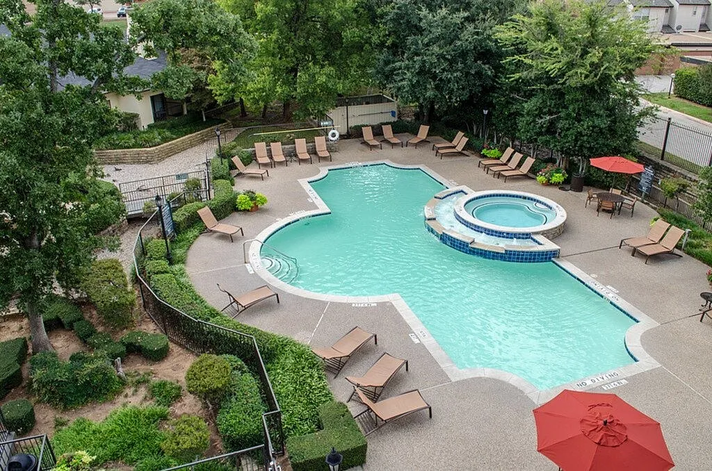Inviting aerial view of a luxury apartment complex pool area, featuring a uniquely shaped pool with adjacent hot tub, surrounded by lush greenery and stylish lounge chairs.