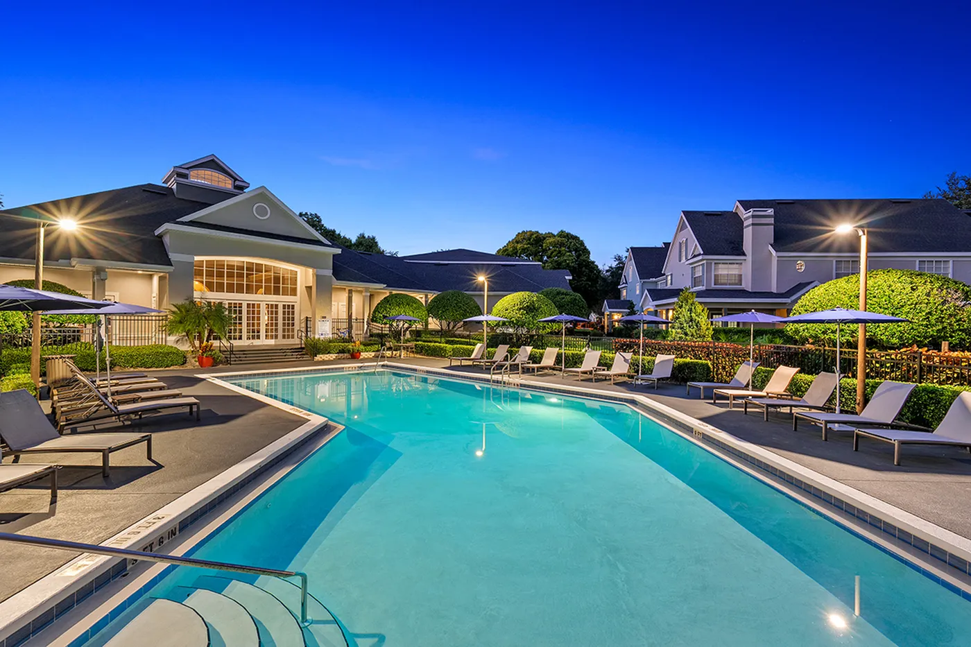 Elegant apartment complex with sparkling swimming pool, surrounded by lush greenery and stylish lounge chairs under a serene evening sky.
