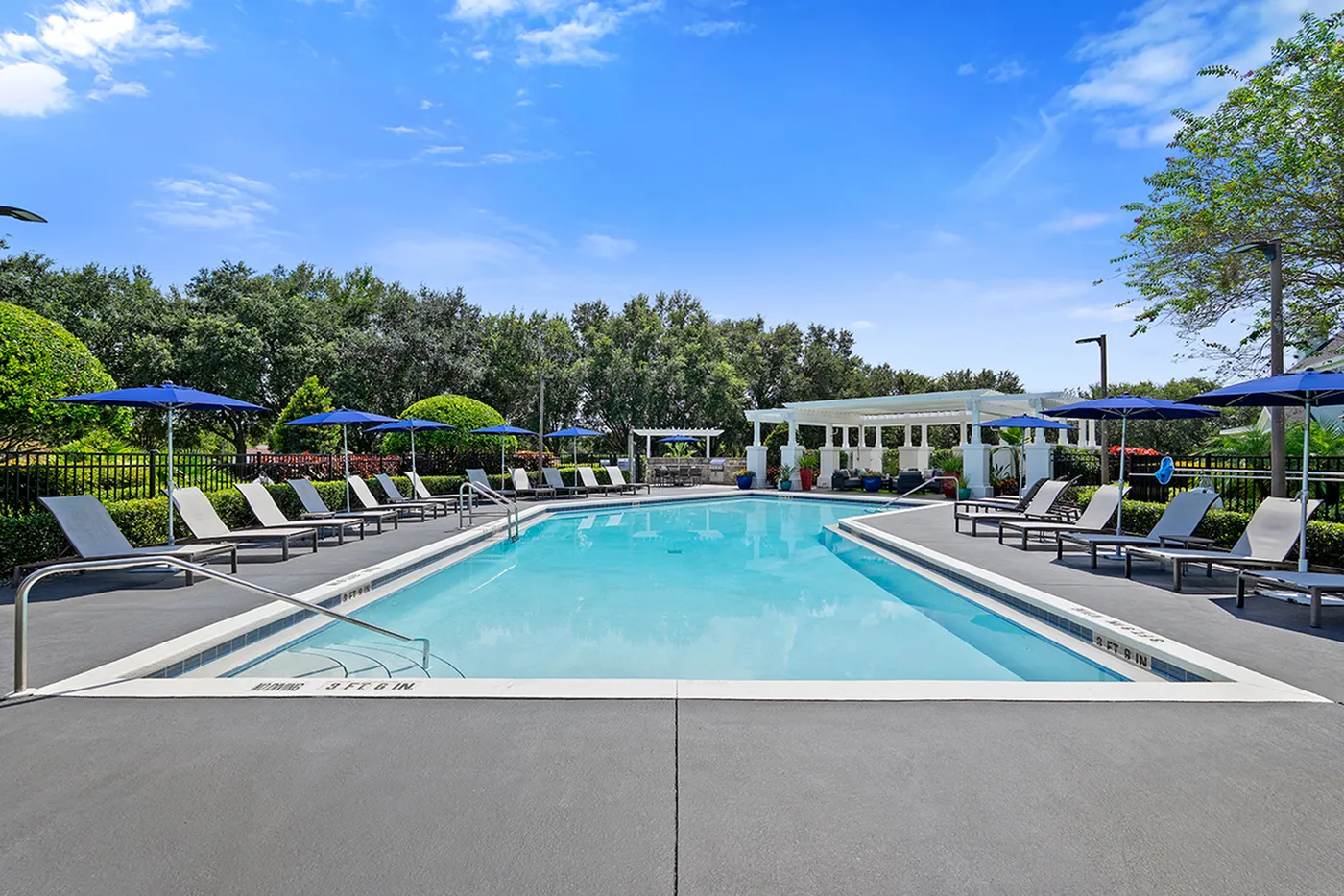 Luxurious outdoor swimming pool area with comfortable lounge chairs, blue umbrellas, and lush greenery under a vibrant blue sky.