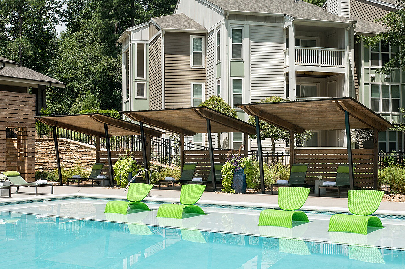Modern apartment community with a refreshing pool and stylish green lounge chairs under sleek wooden cabanas, surrounded by lush landscaping.