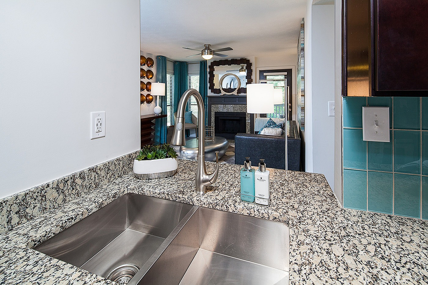 Modern kitchen view with sleek granite countertops, stainless steel sink, and bright living room in the background featuring elegant decor and natural lighting.