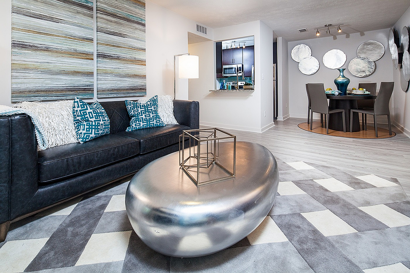 Modern living room with chic black sofa, geometric cushions, and a sleek silver coffee table. Open kitchen and elegant dining area with stylish decor complete the contemporary look.