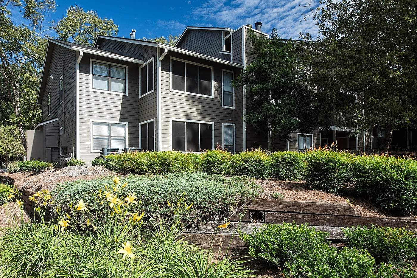 Charming townhouse with modern gray exterior surrounded by lush greenery and beautiful landscaping on a sunny day.