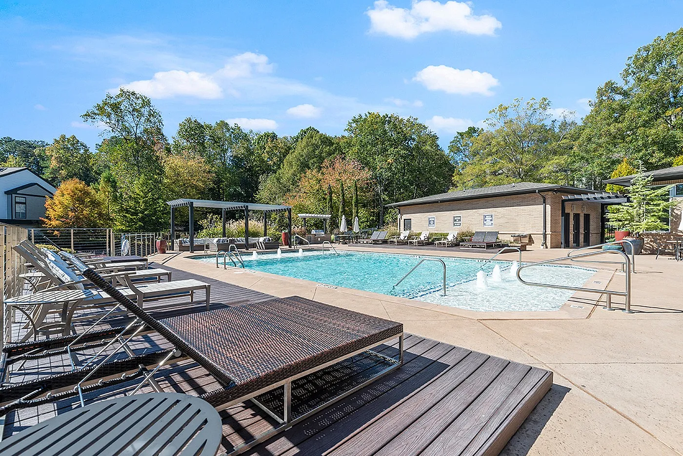 Luxurious outdoor swimming pool area with modern lounge chairs, surrounded by lush green trees and clear blue skies.