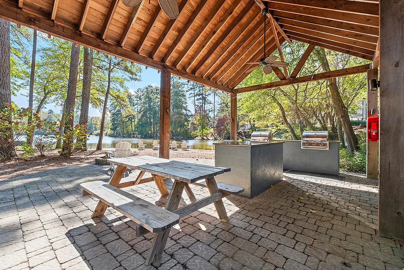 Charming outdoor pavilion with rustic wooden picnic table and modern grills, overlooking a serene lake surrounded by lush trees.