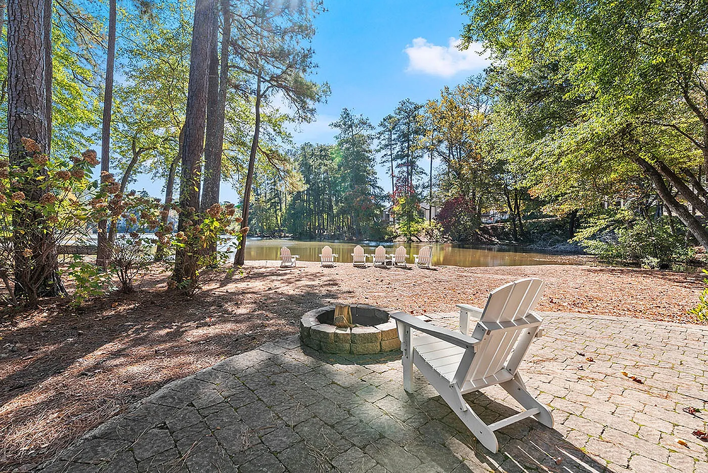 Serene lakeside view with cozy Adirondack chairs, surrounded by lush trees and a picturesque firepit, perfect for relaxation and nature appreciation.