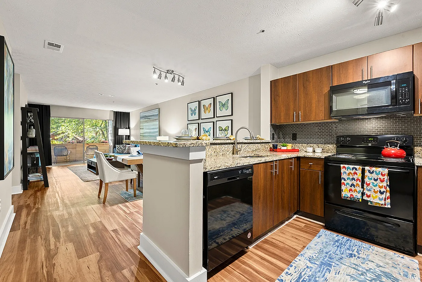 Modern open-concept kitchen and living area with sleek wooden cabinetry, granite countertops, and vibrant decor, leading to a bright seating area with large windows overlooking greenery.