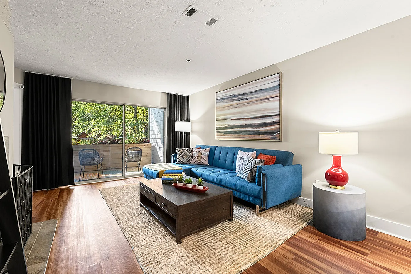 Elegant living room with a stylish blue sofa, modern artwork, and floor-to-ceiling windows opening to a lush balcony view. Perfect blend of comfort and style.