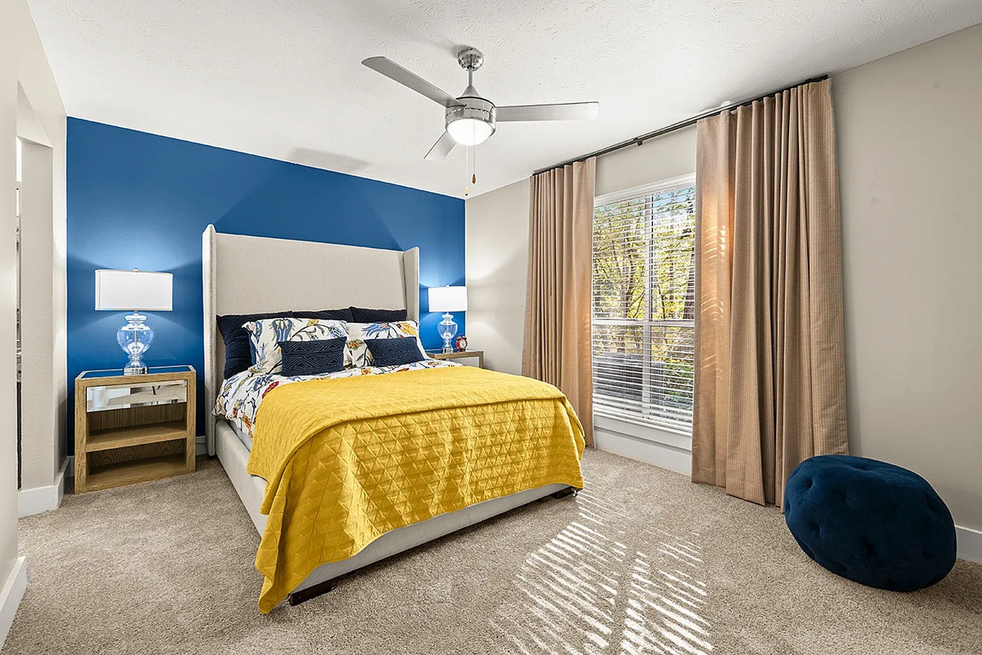 Bright, elegant bedroom featuring a plush bed with vibrant yellow bedding, blue accent wall, and large sunlit window, perfect for a cozy and inviting atmosphere.