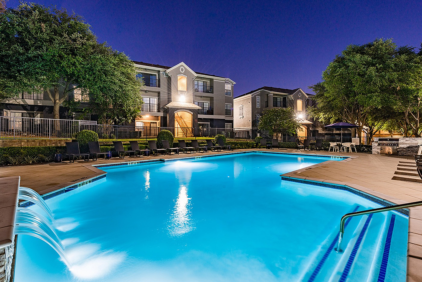Luxurious apartment complex with a sparkling pool illuminated at night, surrounded by lush greenery and elegant architecture.