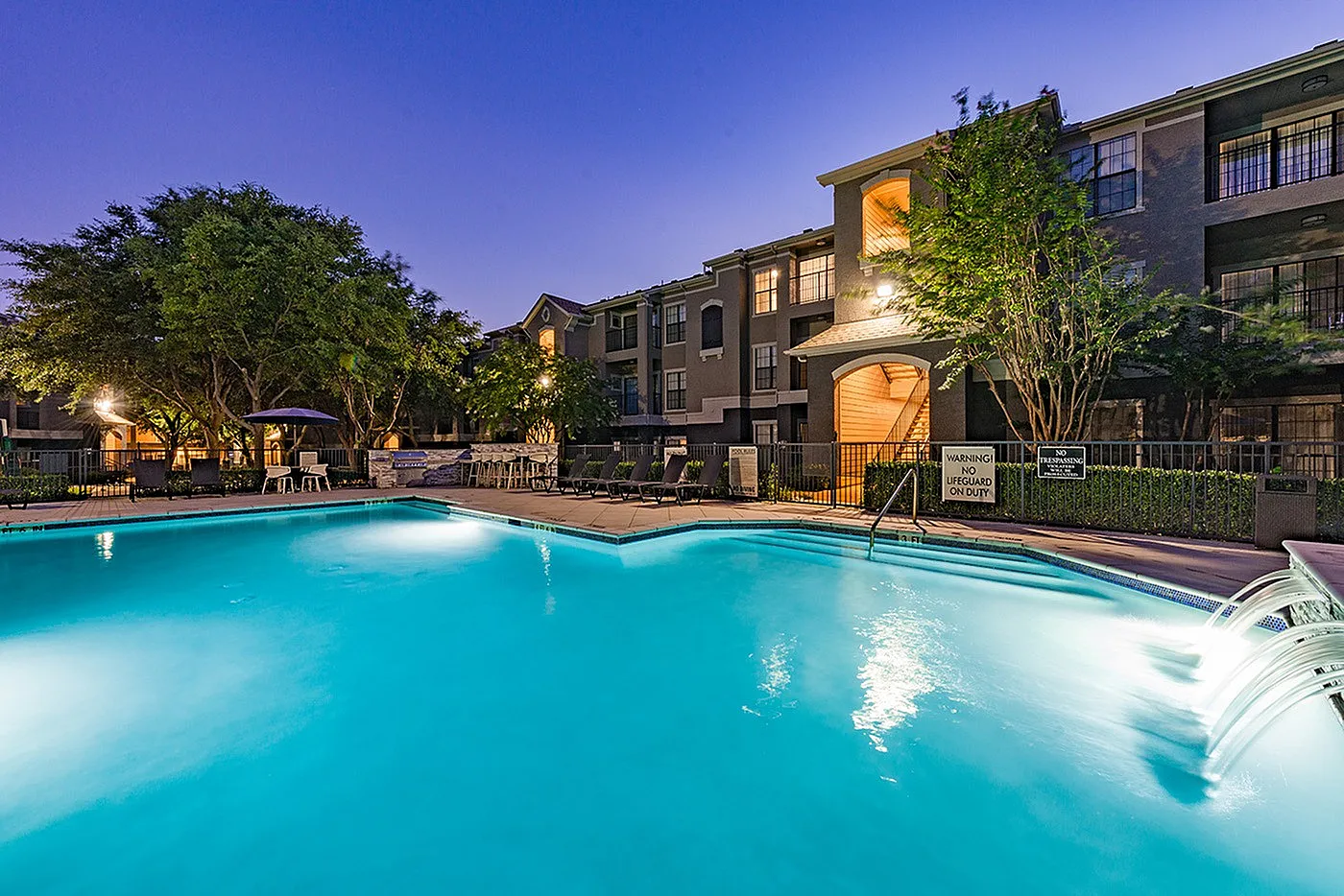 Luxurious apartment complex at dusk featuring a shimmering swimming pool surrounded by lush greenery and inviting lounge chairs.