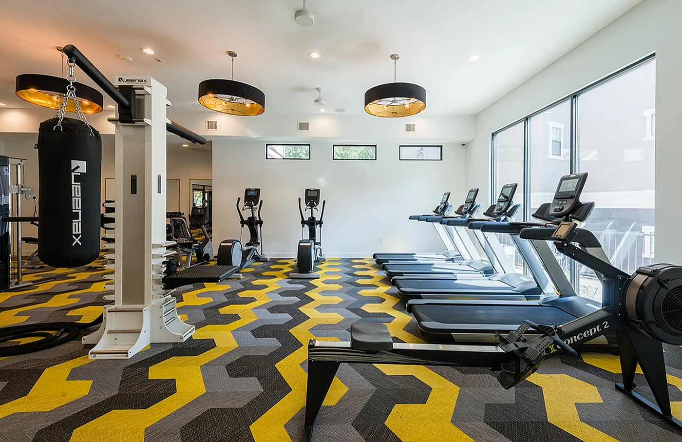 Modern gym with sleek treadmills, rowing machine, and punching bag on vibrant geometric flooring, illuminated by stylish overhead lights.