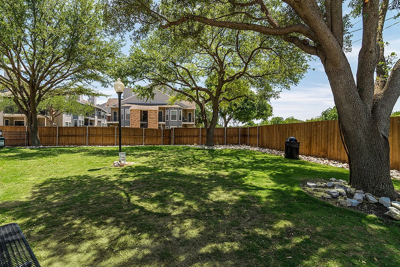 Inviting apartment community outdoor area with lush green lawn, mature trees, and charming wooden fence under a clear blue sky.
