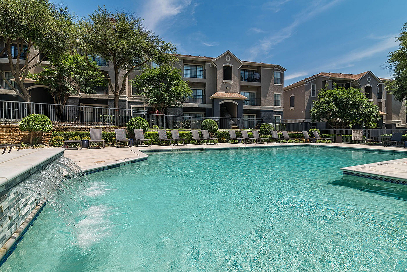 Charming apartment complex with lush landscaping, featuring a stunning outdoor pool and relaxing lounge chairs under the bright blue sky.