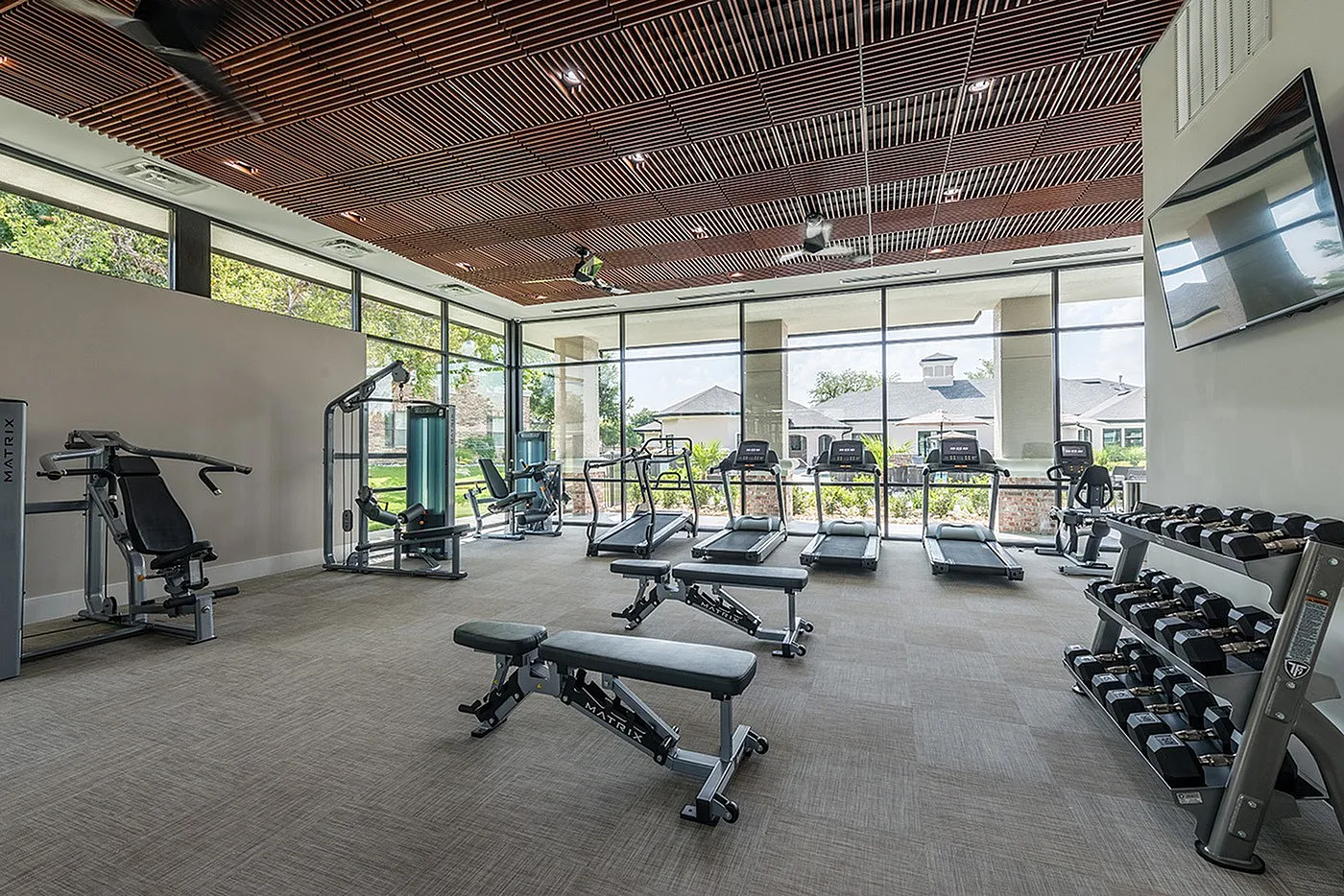 Modern fitness center with state-of-the-art gym equipment, large windows offering natural light, and a sleek wooden ceiling design.