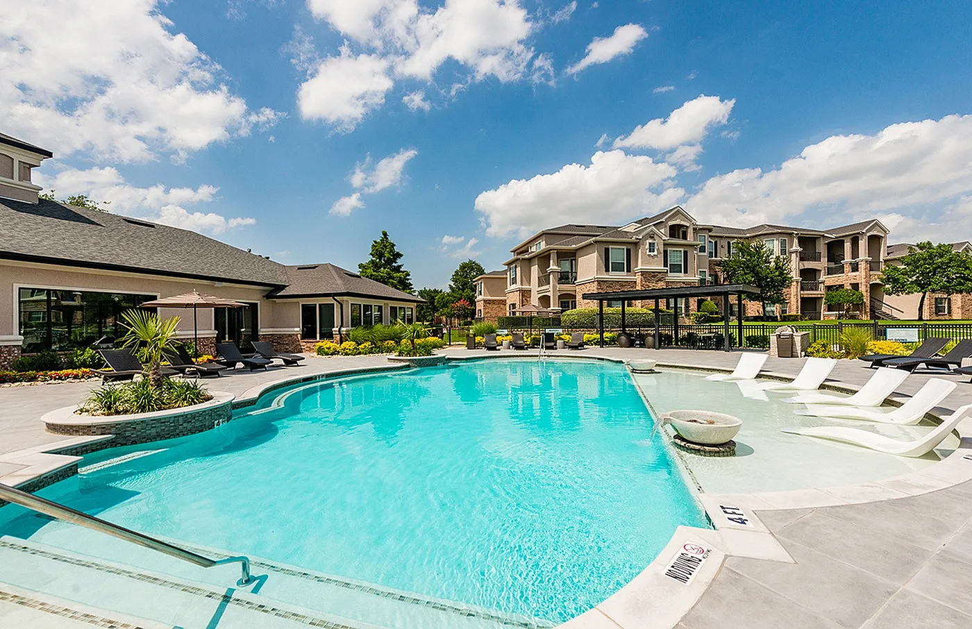 Luxurious apartment with a sparkling swimming pool, poolside loungers, and lush landscaping under a bright blue sky.