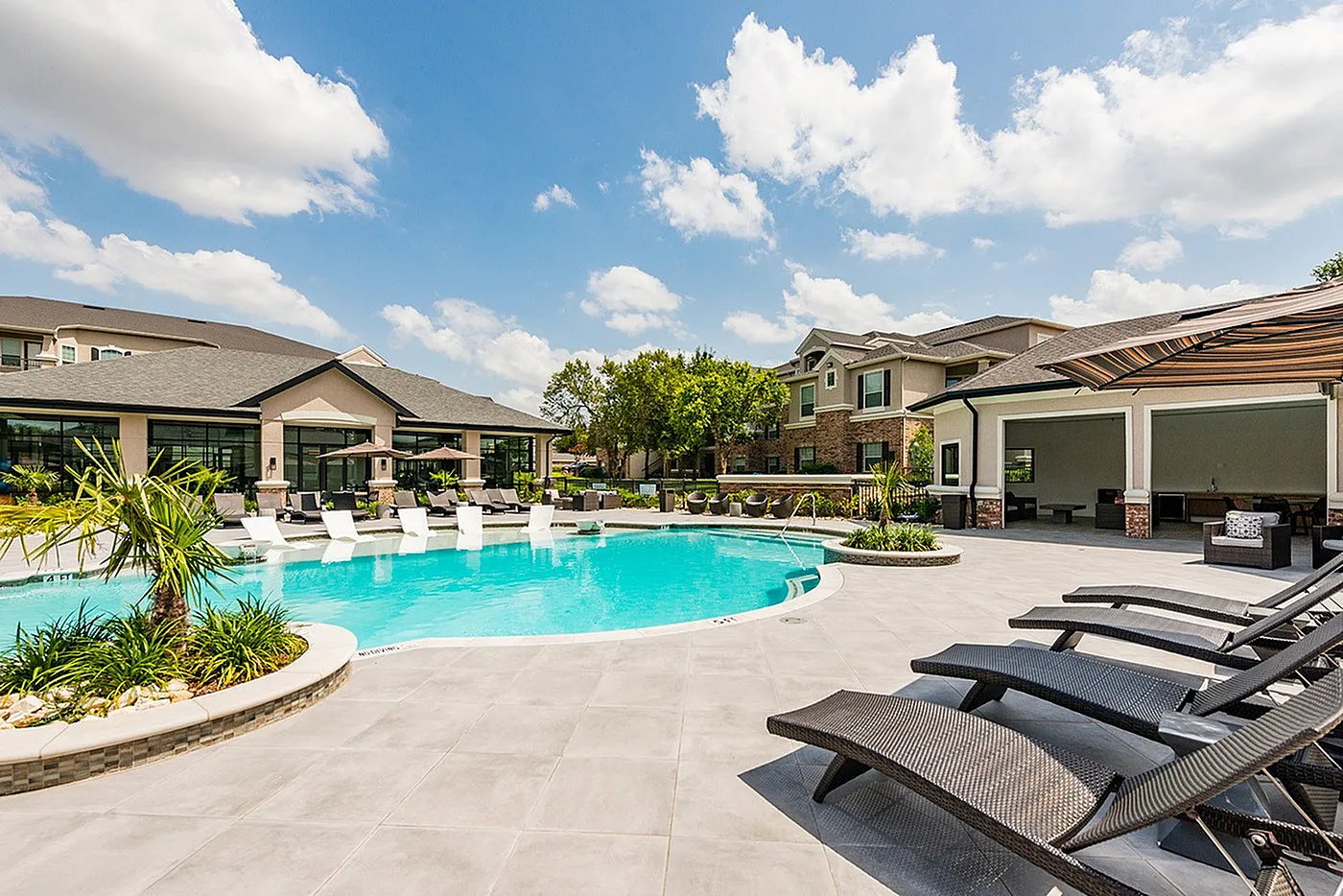 Luxurious apartment complex featuring a sparkling outdoor pool surrounded by elegant lounge seating, lush landscaping, and modern architecture under a bright blue sky.