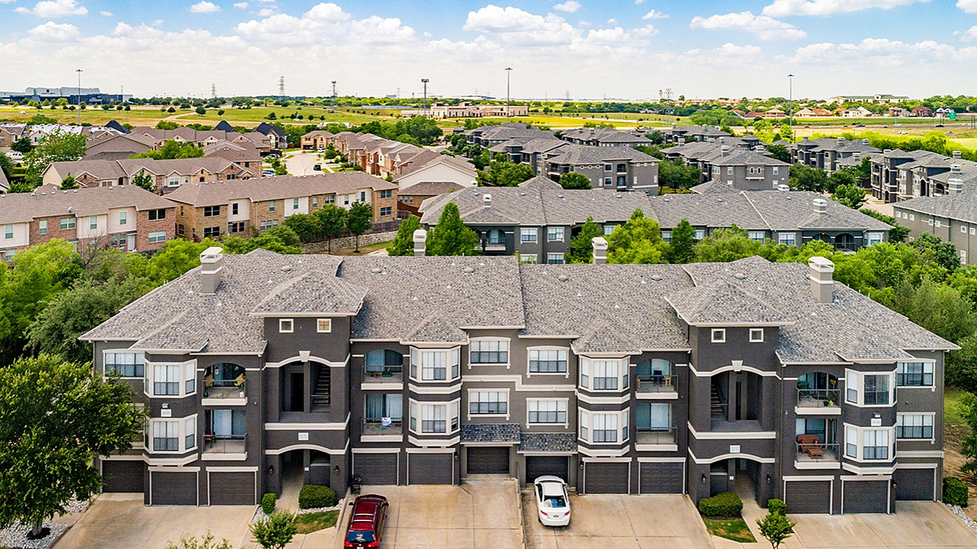 Picturesque aerial view of modern apartment complex with elegant architecture, lush green surroundings, and ample parking spaces, offering a serene suburban lifestyle.