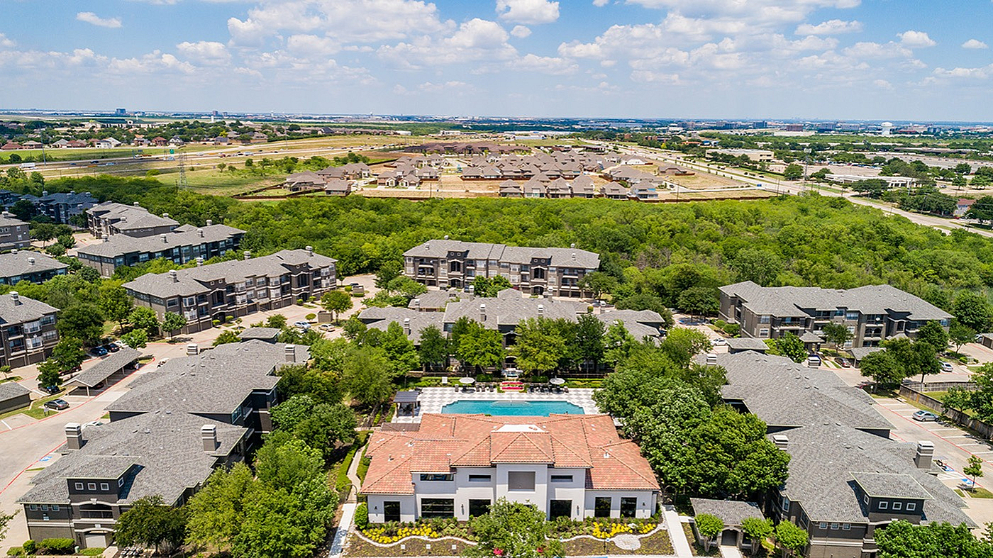 Aerial view of a lush apartment community with modern buildings, centered around a sparkling pool and surrounded by green spaces, offering tranquil suburban living.