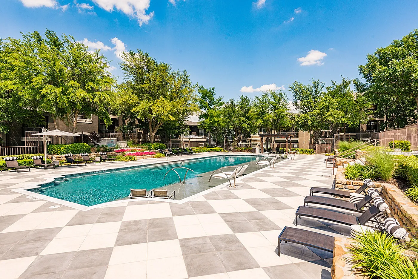Luxurious apartment complex pool area featuring a sparkling swimming pool, elegant sun loungers, lush greenery, and a stylish checkered patio under a clear blue sky.