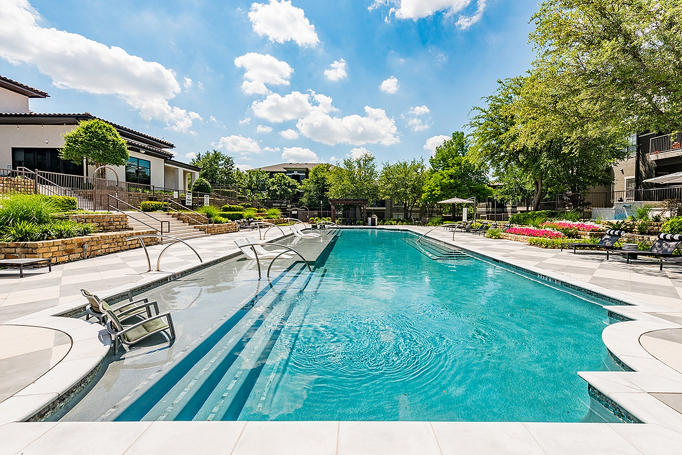 Luxurious outdoor pool area with modern lounge chairs, surrounded by lush greenery and vibrant floral landscaping under a bright blue sky.