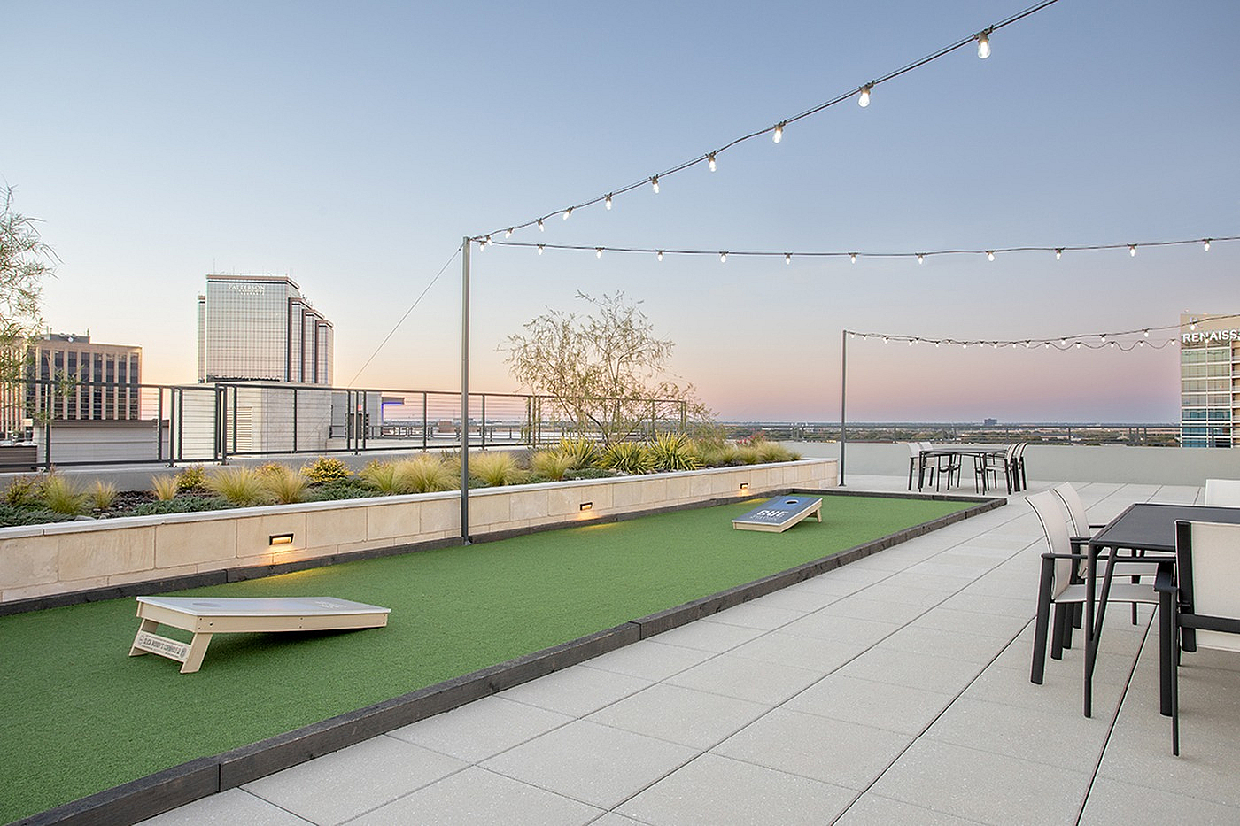 Modern rooftop terrace with comfortable seating, cornhole game, and ambient string lights against a vibrant cityscape.