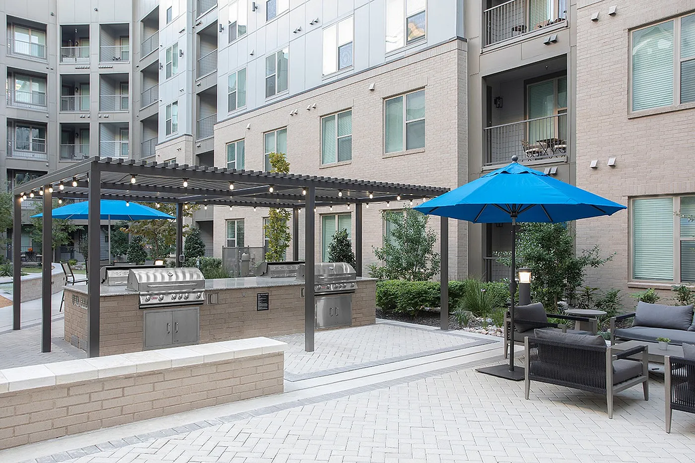 Modern apartment courtyard with sleek grilling station, cozy seating under blue umbrellas, and elegant string lights.