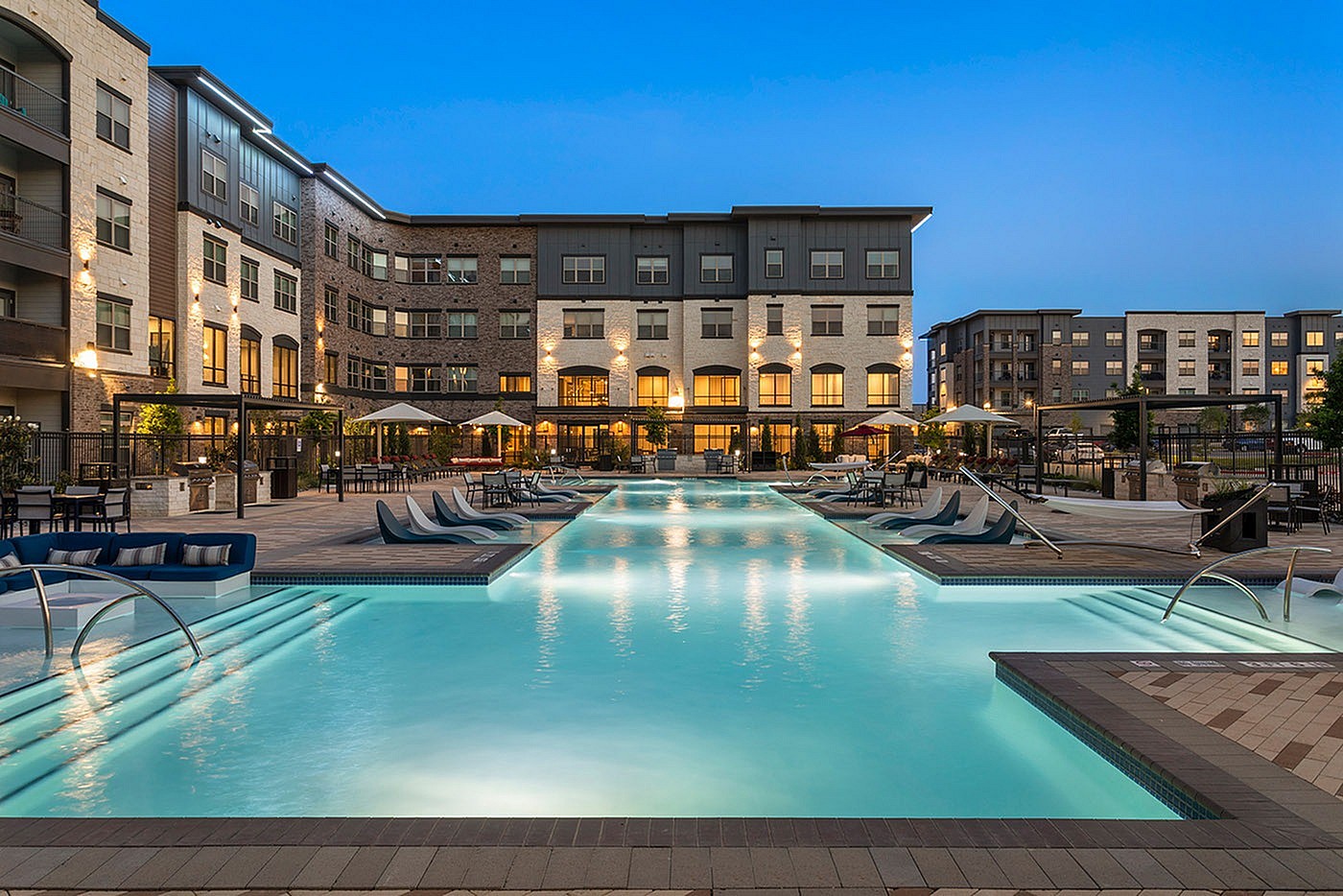 Luxurious apartment complex with a sparkling outdoor pool featuring stylish lounge chairs and illuminated modern architecture.