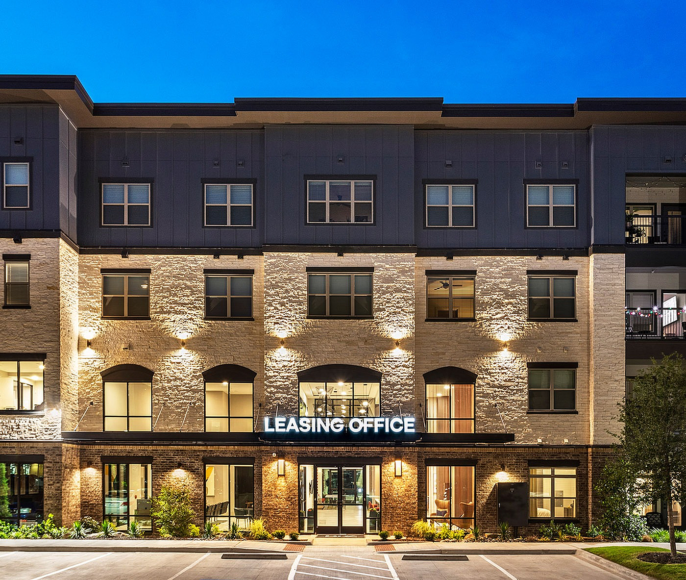 Elegant modern leasing office exterior, warmly lit at dusk, featuring pristine brickwork and large windows.