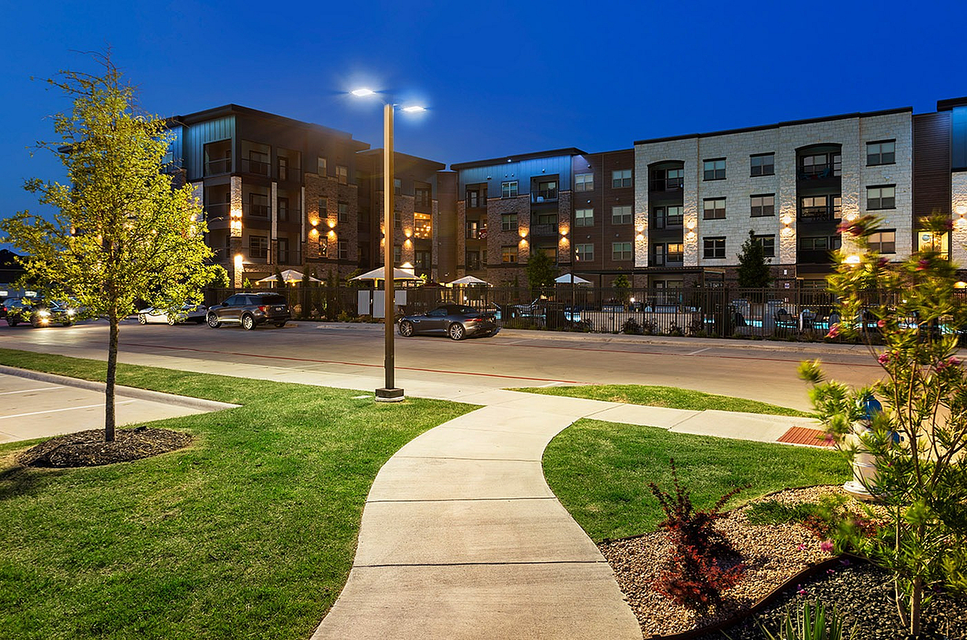 Elegant apartment complex illuminated at night, showcasing modern architecture and landscaped surroundings with a welcoming pathway.