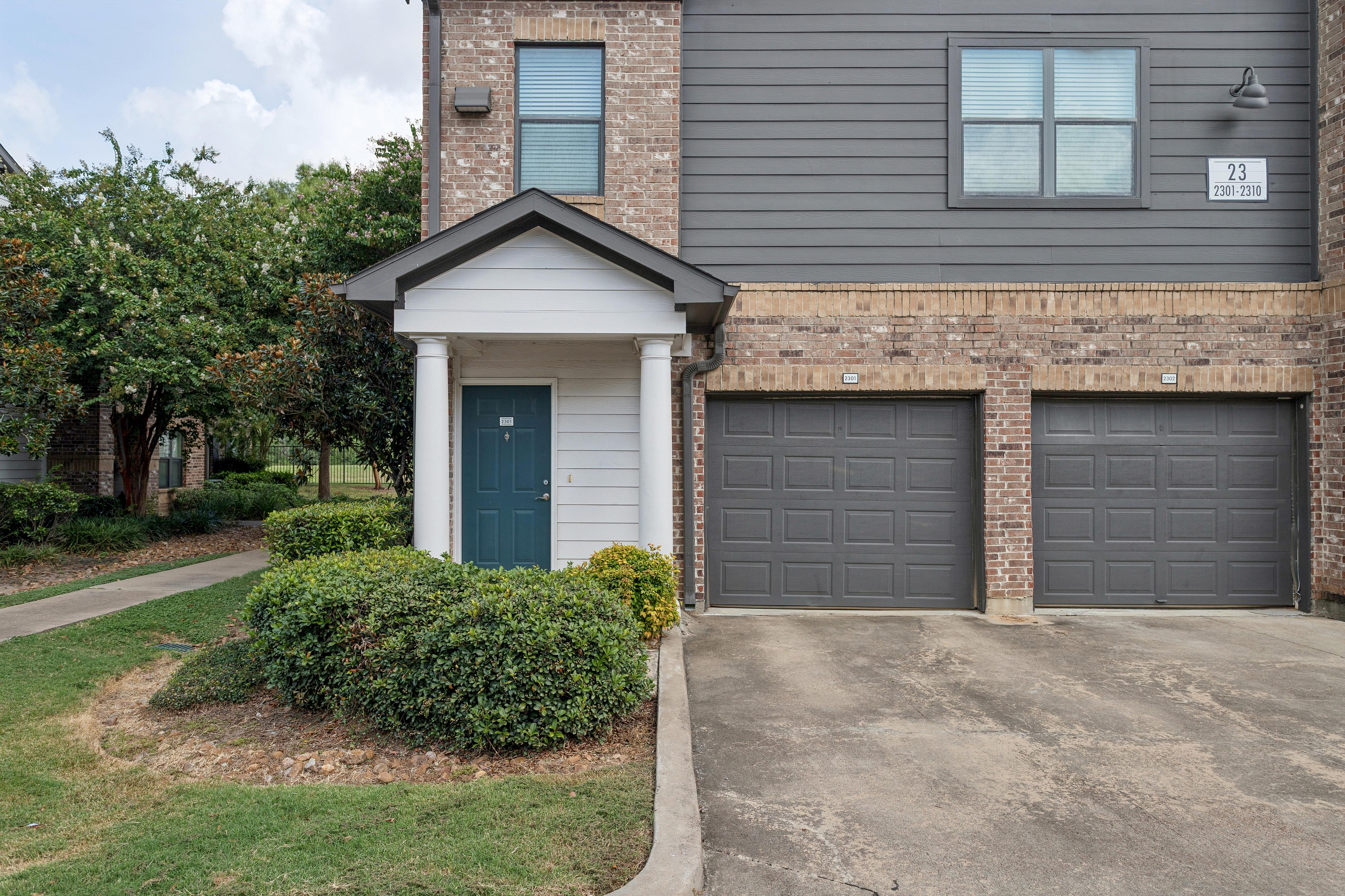 Charming apartment exterior with a stylish blue door, dual garages, and lush greenery, offering a welcoming and modern appearance.
