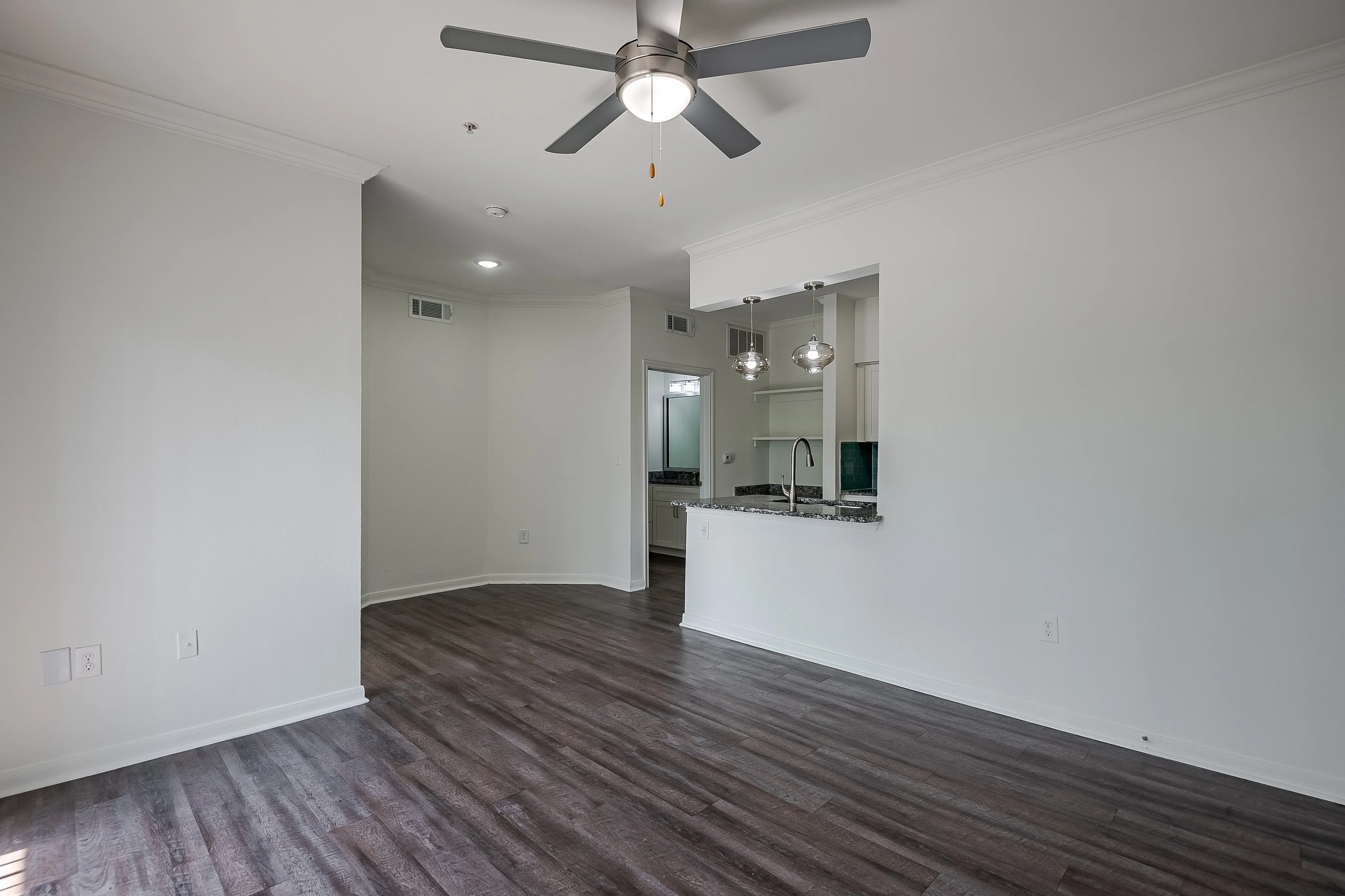 Chic living space with sleek hardwood floors, modern ceiling fan, and open kitchen area featuring stylish pendant lighting.