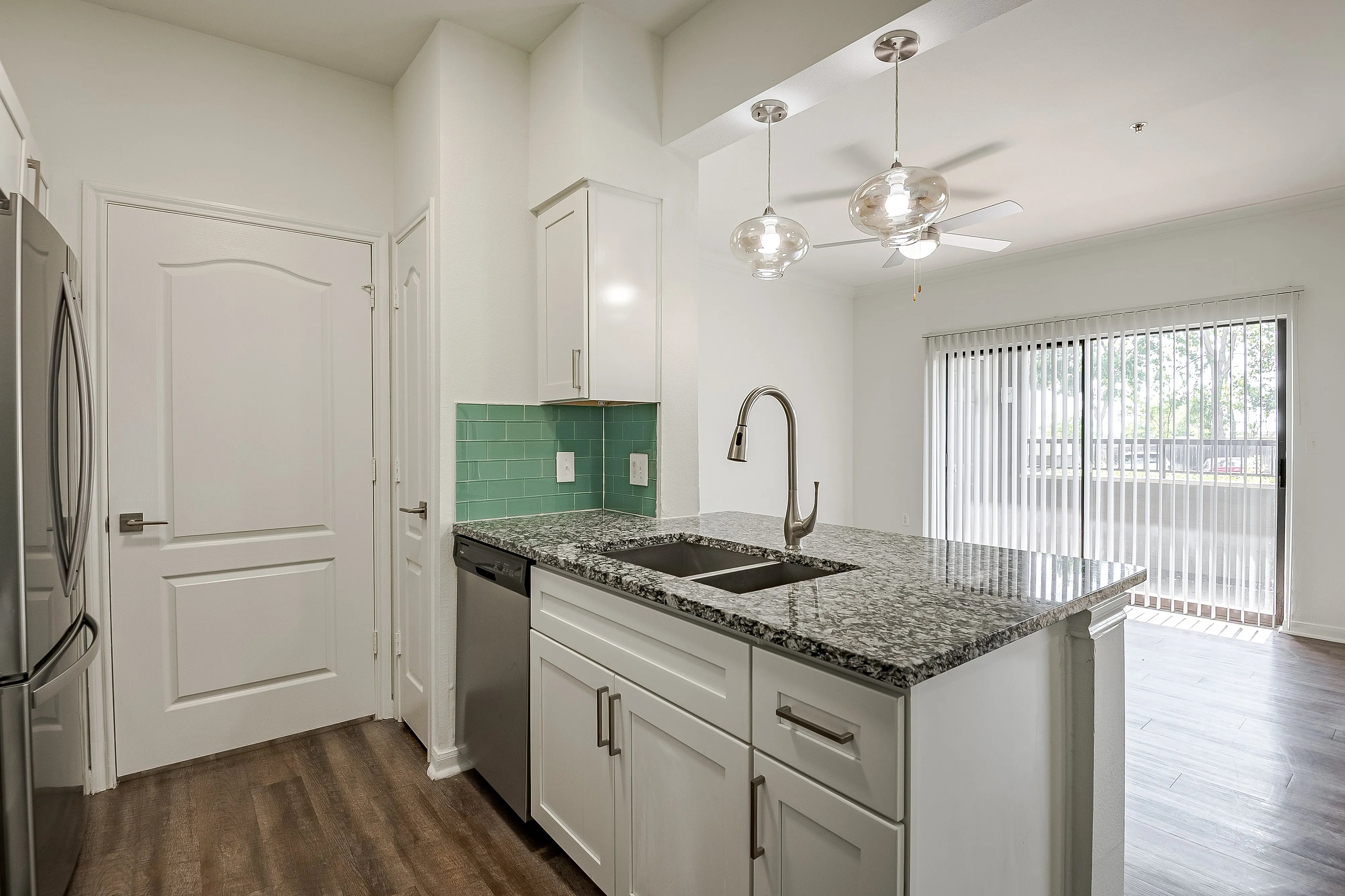Modern kitchen with elegant granite countertops, sleek stainless steel appliances, and chic pendant lighting, leading to a bright living area with large glass doors.