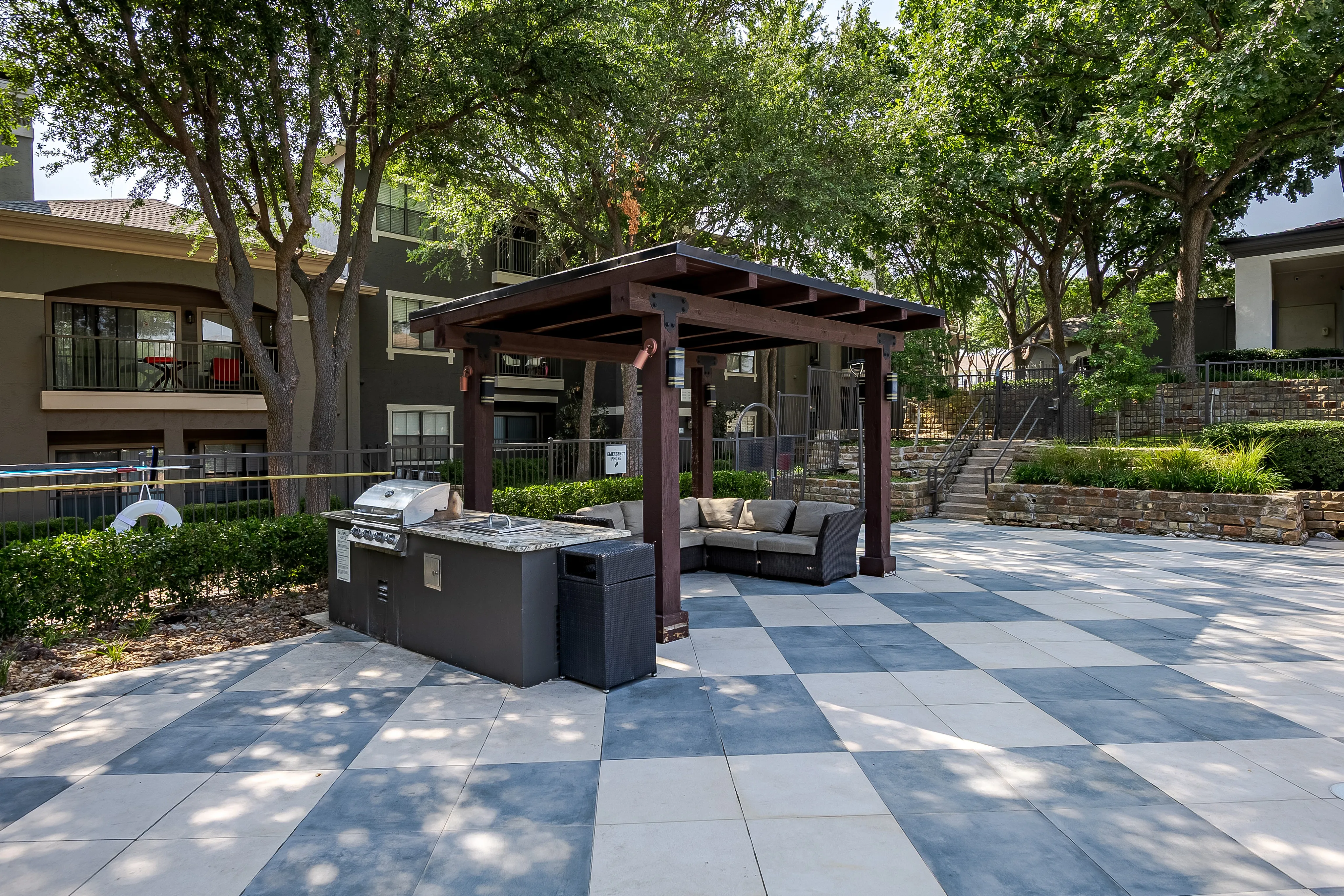 Charming outdoor patio with BBQ grill, shaded pergola, and cozy seating area in a lush apartment community setting.
