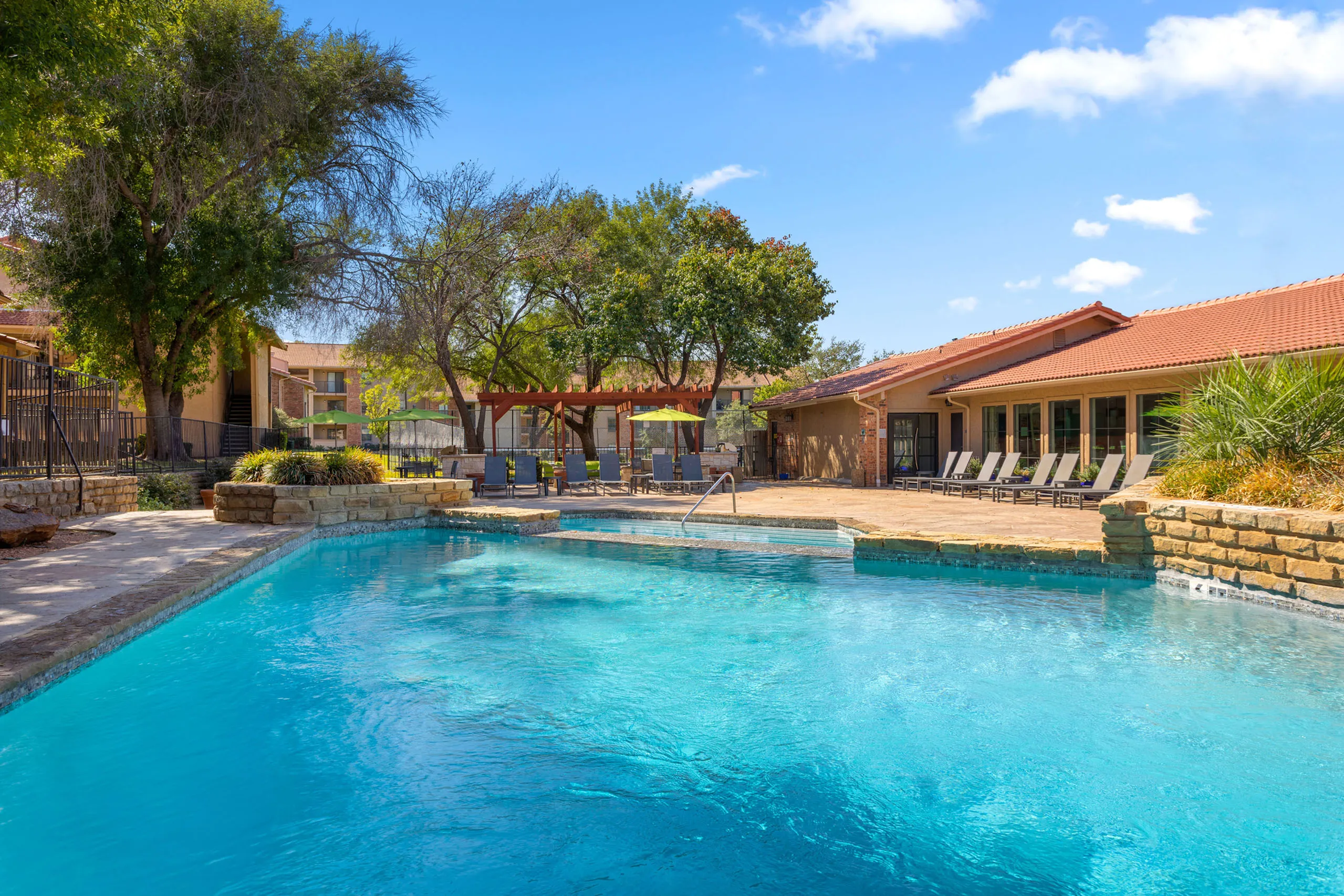 Inviting outdoor pool area with sun loungers, surrounded by lush greenery and a charming clubhouse.