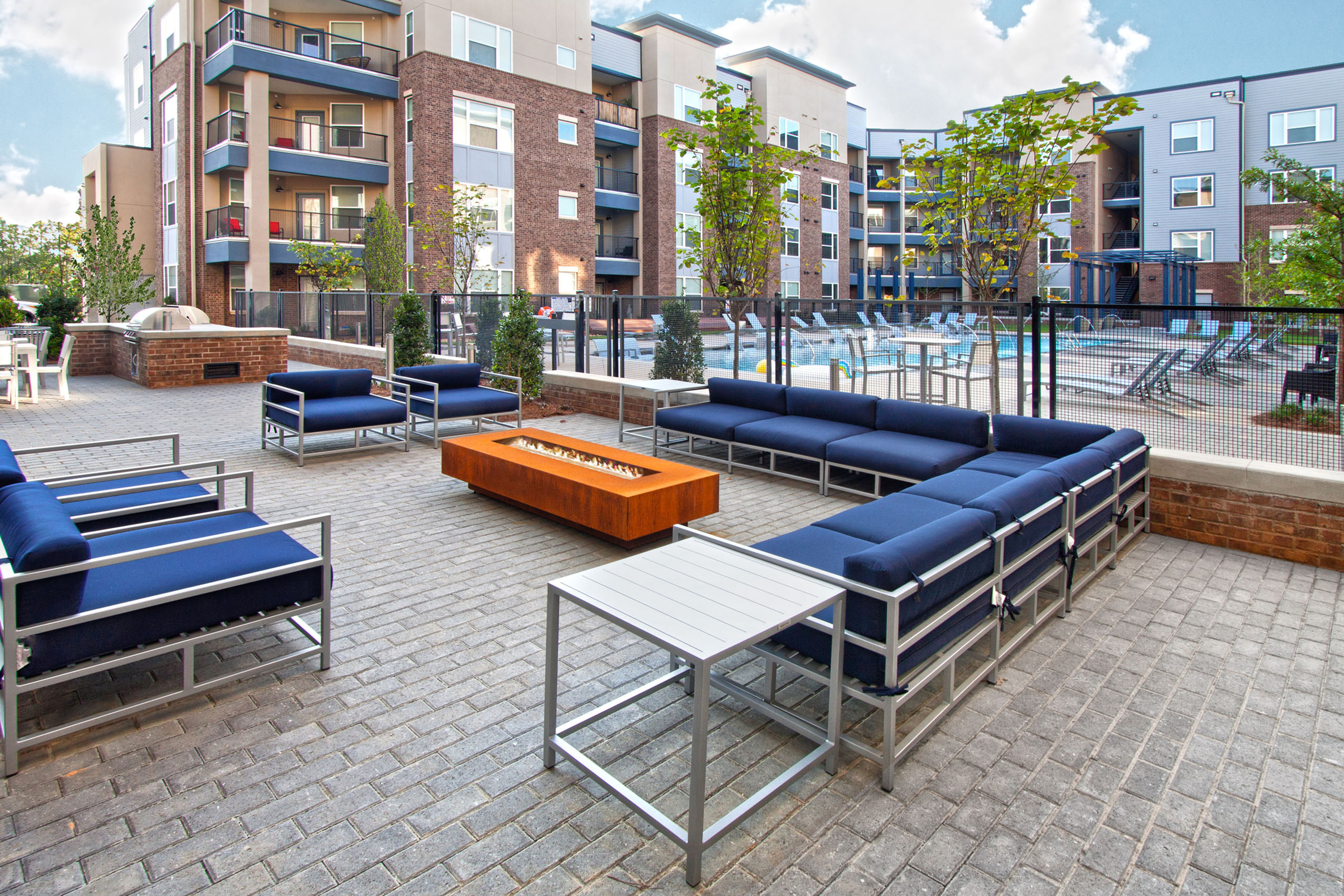 Modern apartment complex showcases a vibrant outdoor lounge with stylish seating and a cozy fire pit, adjacent to a sparkling pool and surrounded by lush greenery.