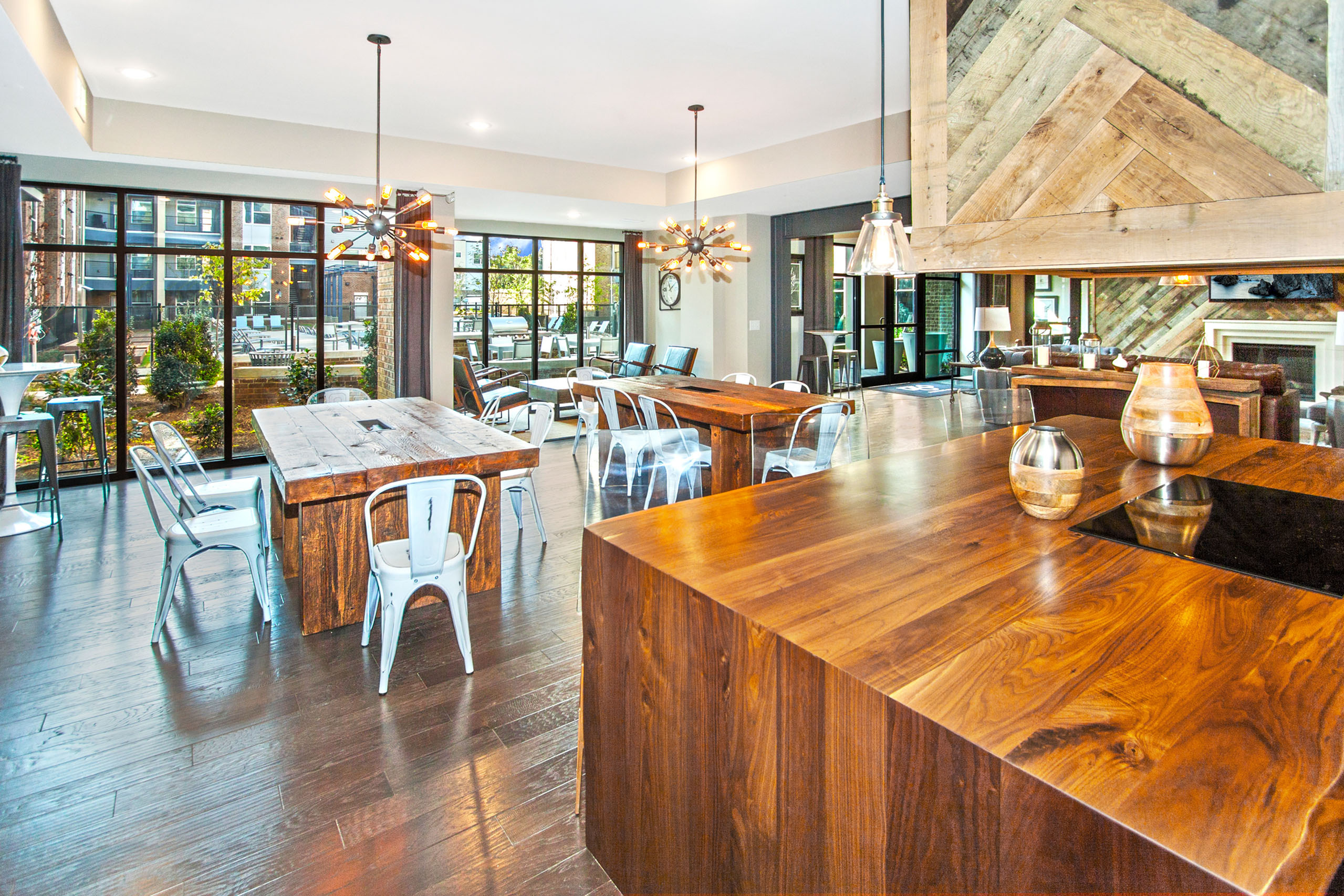 Chic communal space with rustic wooden tables, modern chairs, and stylish pendant lighting, surrounded by floor-to-ceiling windows offering a scenic view.
