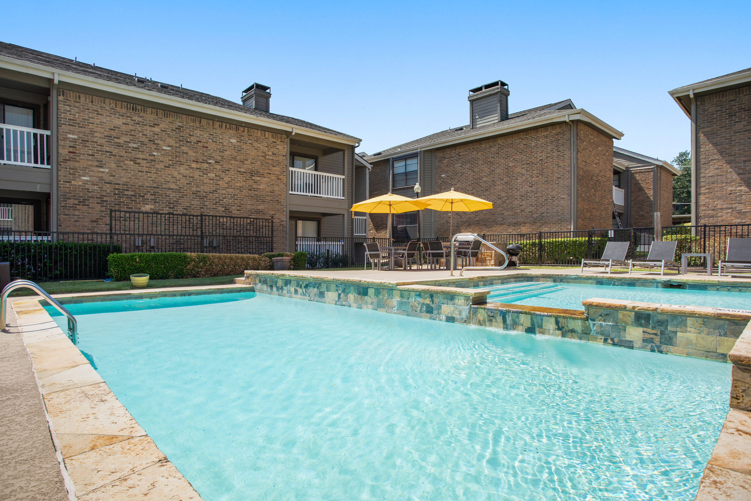 Inviting community pool area with sparkling waters, surrounded by elegant brick apartment exteriors and vibrant yellow umbrellas.