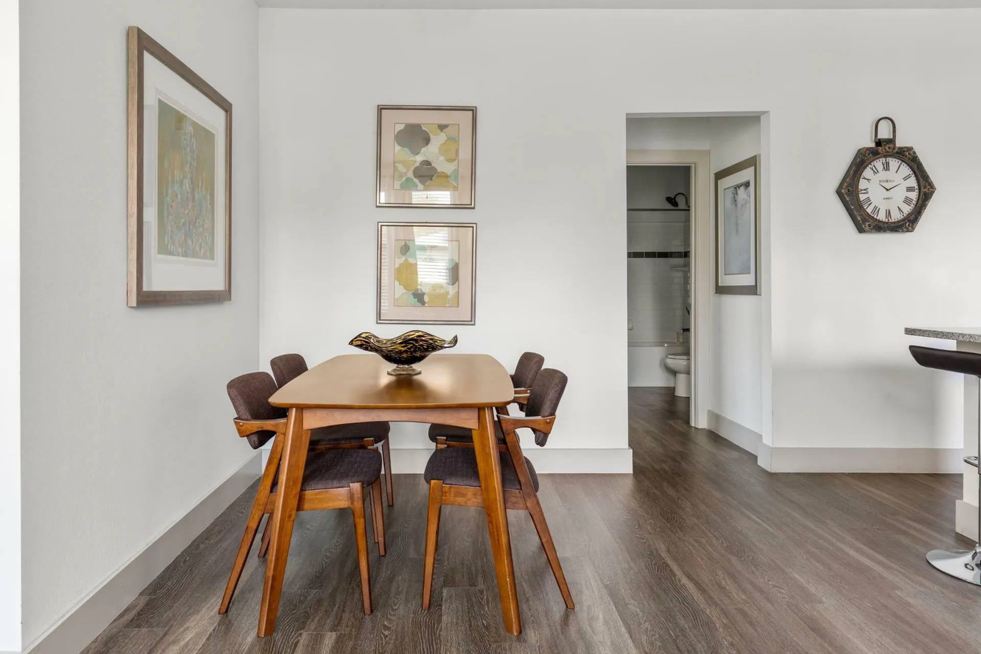 Elegant dining area with modern wooden table and chairs, art-adorned walls, leading into a sleek bathroom, perfect for stylish living.