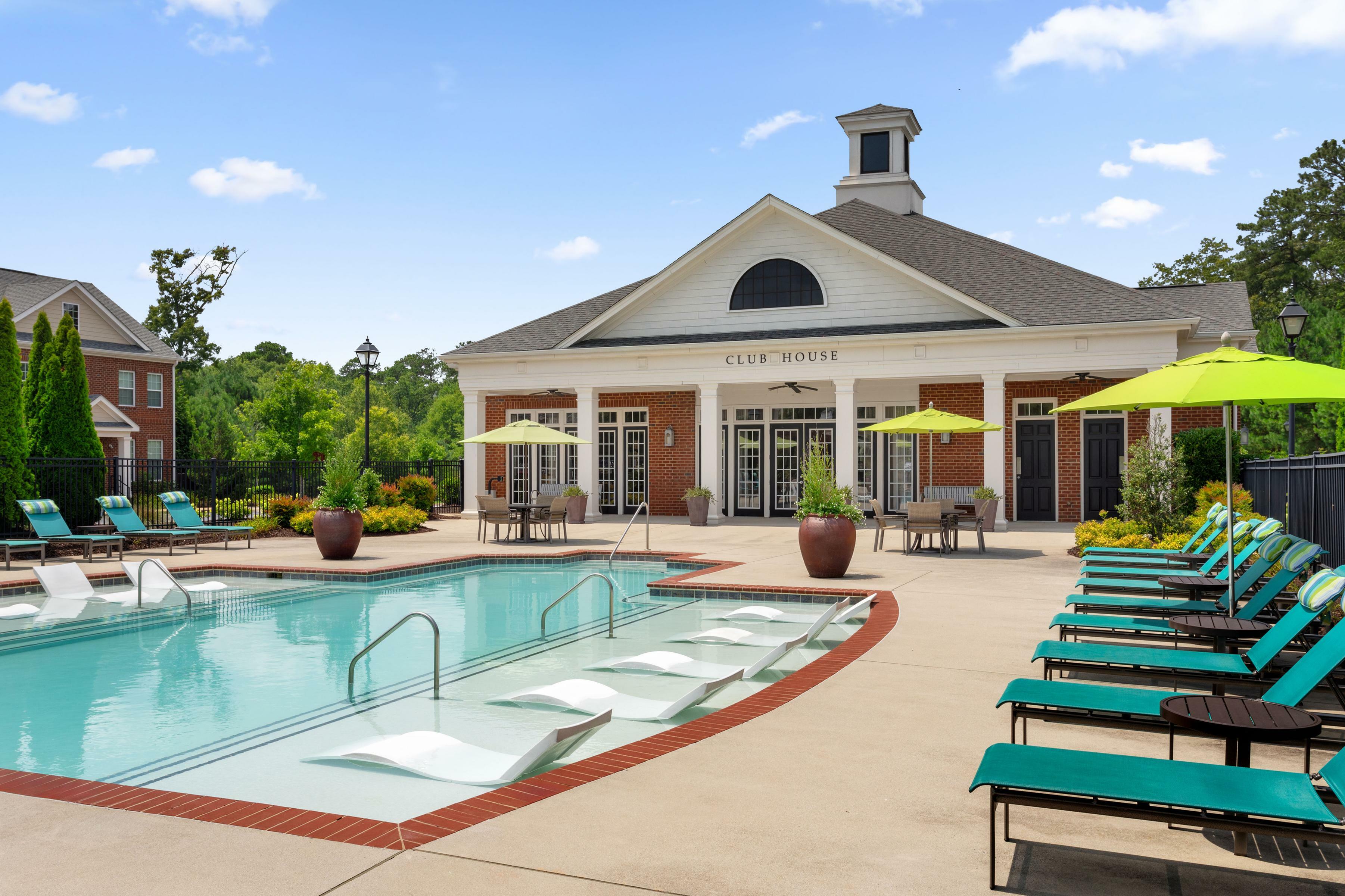 Elegant clubhouse with inviting swimming pool, surrounded by vibrant green sun loungers and umbrellas, set in a serene landscaped community.