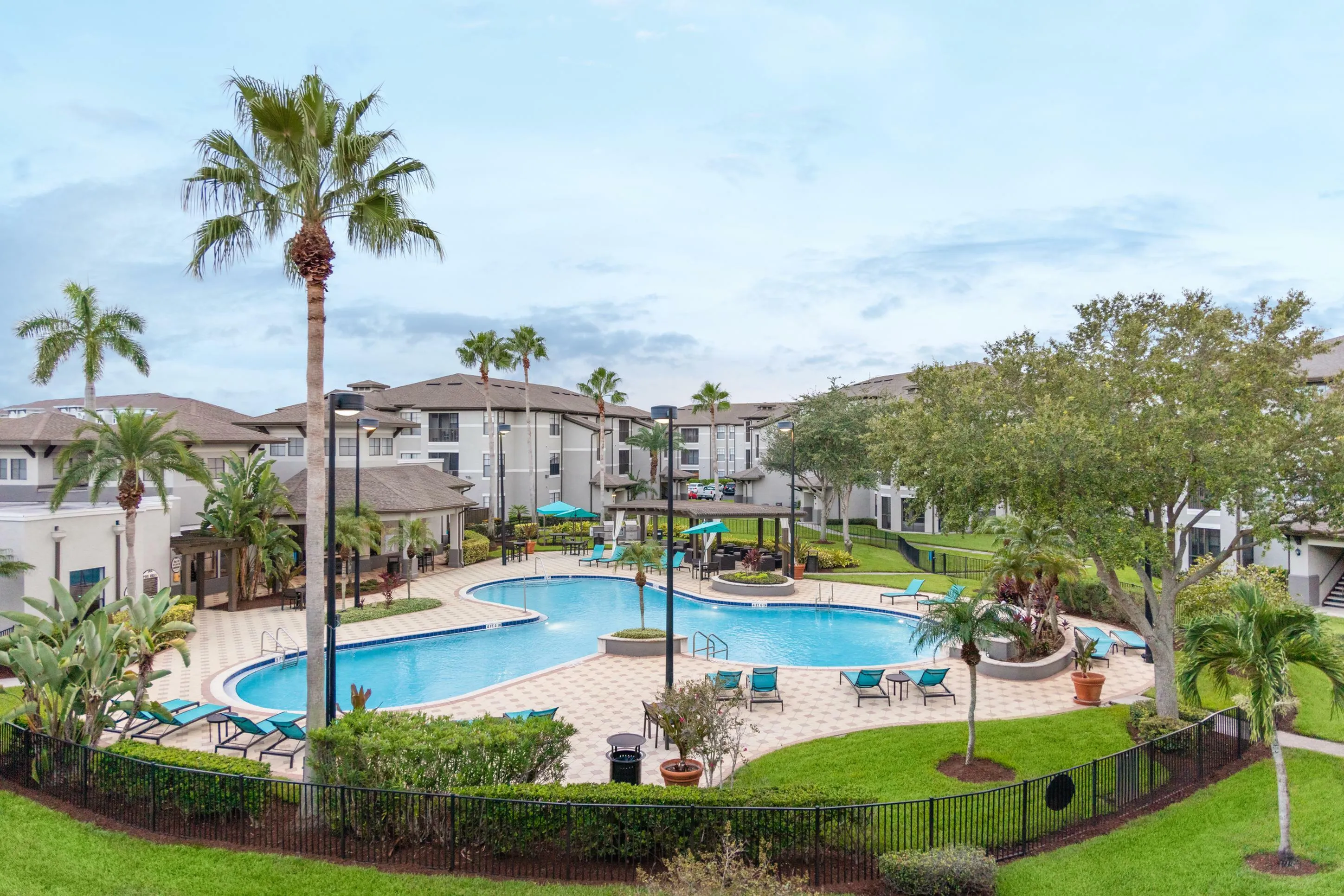 Luxurious apartment complex with resort-style pool, palm trees, and lush landscaping under clear skies.